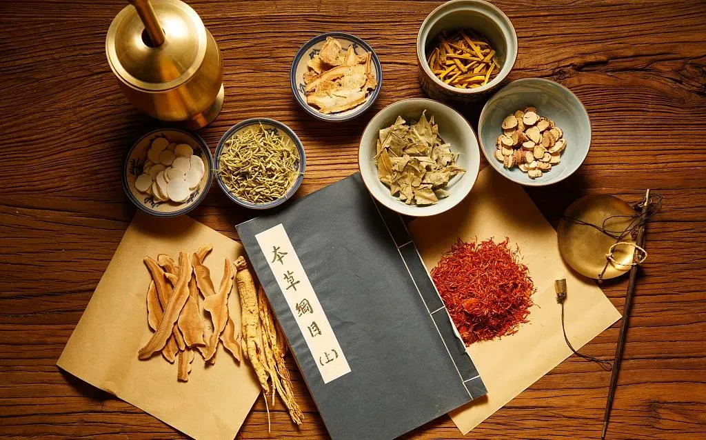 Traditional Chinese medicinal herbs arranged in bowls beside an old herbal compendium